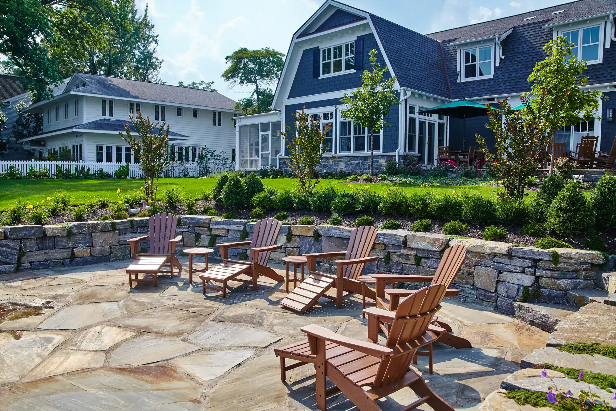 A backyard patio in NY featuring wooden Adirondack chairs around a small table, stone flooring crafted by a masonry contractor Long Island, a low stone wall with shrubs, and a large house with gray siding and white trim in the background.