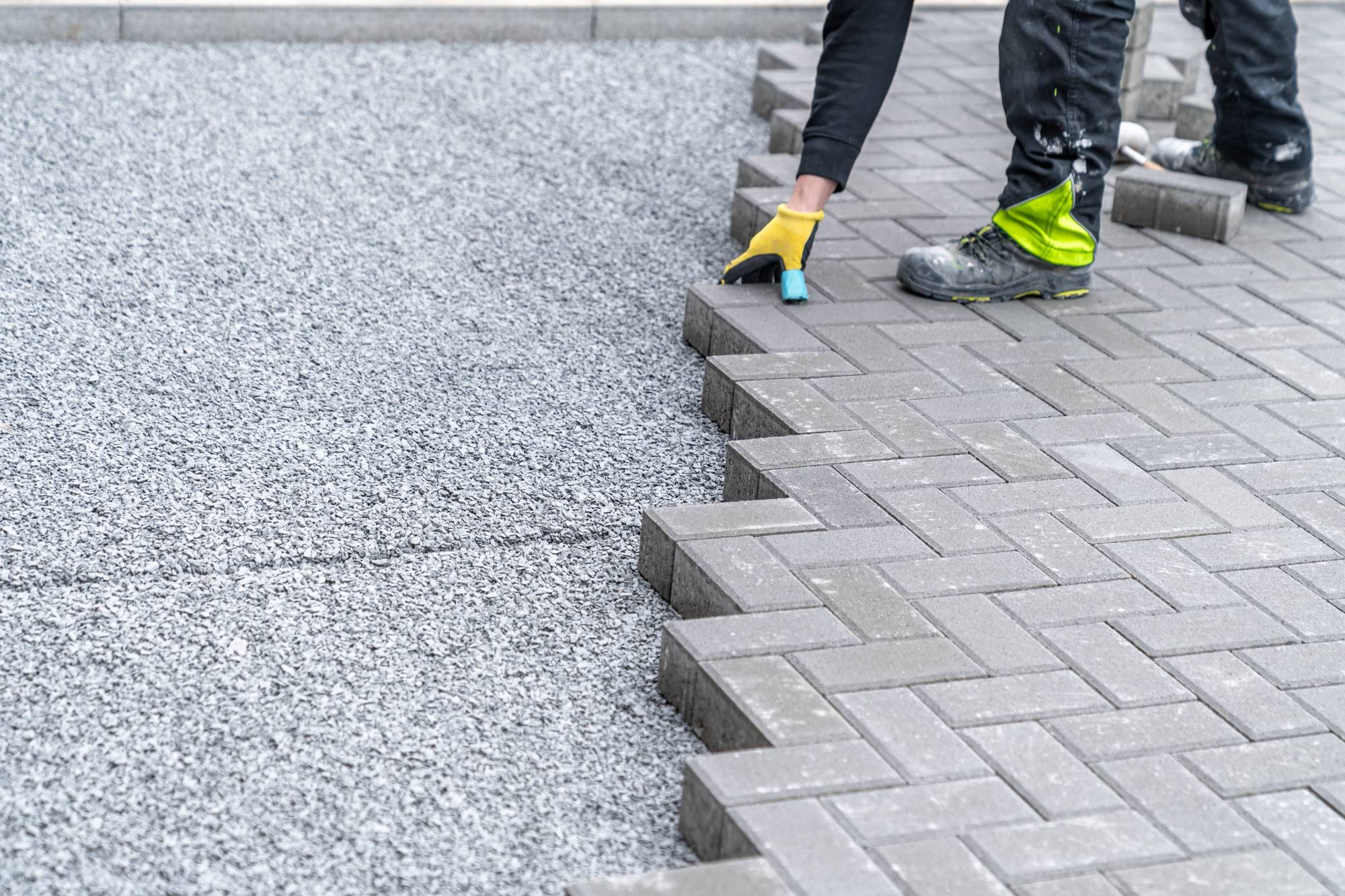 A worker wearing gloves and work boots, from a masonry contractor Long Island, is laying gray rectangular paving stones in a herringbone pattern on a gravel base for a pathway or driveway in NY.